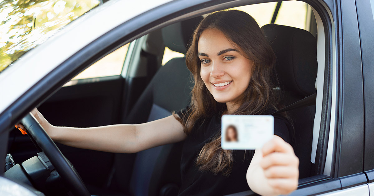 A woman with long brown hair sits in a car's driver's seat, smiling and holding up a driver's license towards the camera, conveying a sense of achievement.