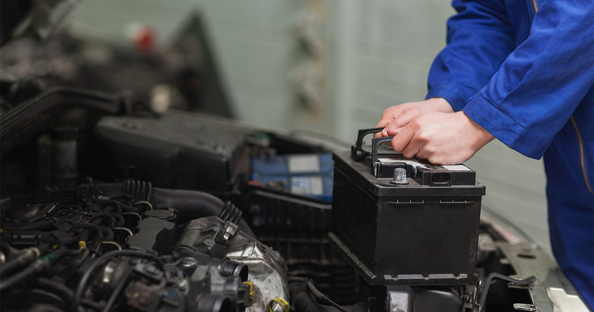 A mechanic in blue coveralls installs a car battery under the hood, focusing on the task. The scene conveys a professional, hands-on approach.