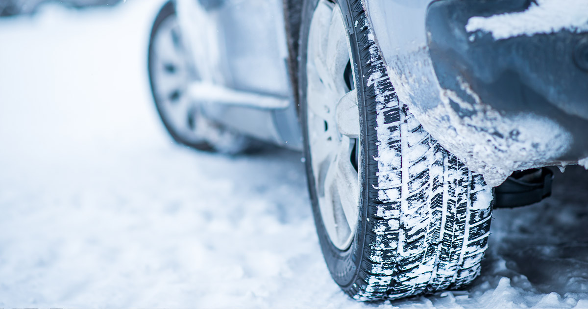 Close-up of a car tire on a snowy road. The tire's tread is clearly visible, surrounded by fresh snow. The scene conveys a sense of winter travel.