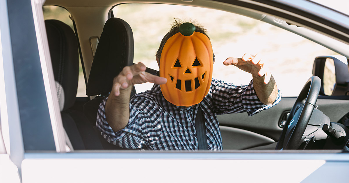 A person in a checkered shirt, wearing a pumpkin mask, playfully reaches out from a car window. The scene is humorous and Halloween-themed.