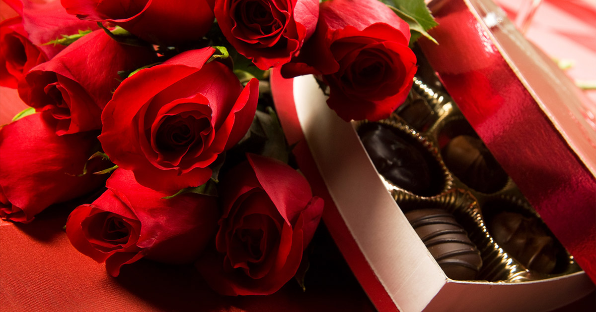 A bouquet of vibrant red roses beside an opened heart-shaped box of assorted chocolates on a red surface, conveying romantic elegance and affection.